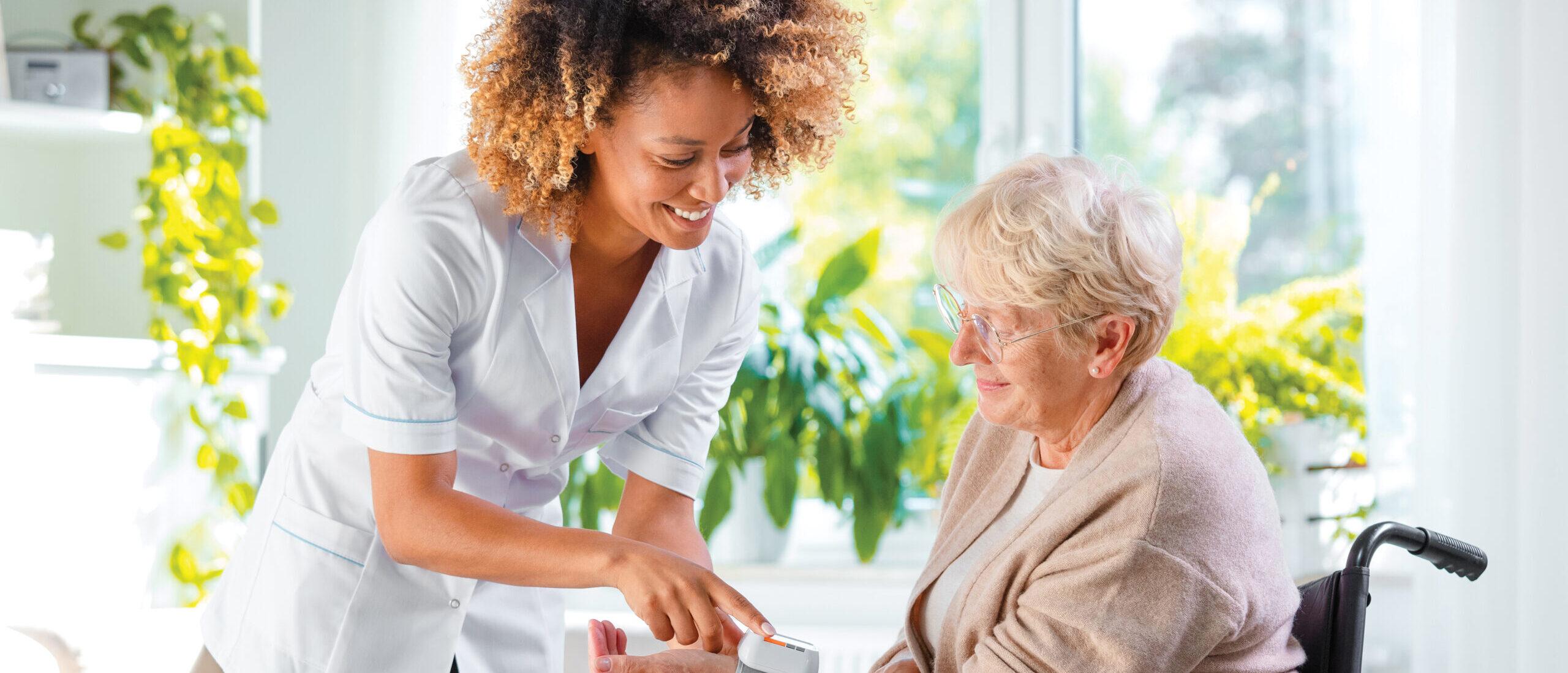 A home nurse checks the blood pressure of a woman in a wheelchair. They both look happy and content.